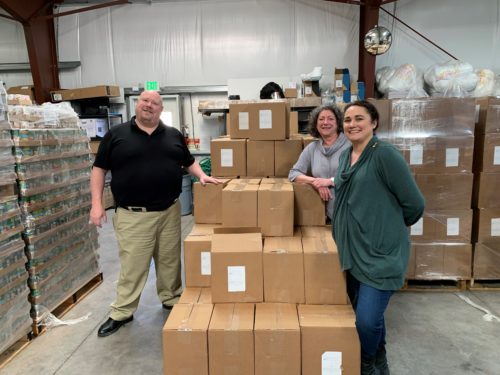 Three people stand in a warehouse with boxes of food